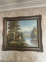 Photo of an ornate framed landscape painting showing a country summer scene with water, trees, and mountains in the background on wallpapered wall
