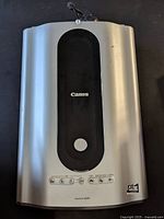 Canon CanoScan 8600F flatbed scanner showing front and top, silver casing with black center panel and a round control button.