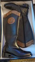 Pair of tall black leather boots with brown trim laid side by side inside box, showing side zipper and embossed logo patch on shaft.