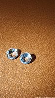 Two natural Brazilian aquamarine gemstones displayed on textured brown background, both round and faceted, light blue color, casting distinct shadows.
