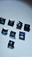 Nine natural blue sapphire princess cut gemstones in deep blue color, each approximately 2.7-3mm in size, shown laid out with light reflections highlighting facets and clarity.