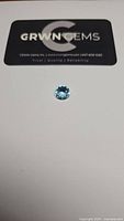 Photo of a round cut blue topaz gemstone placed on a white surface with a black and silver GRWN Gems card above it.