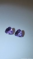 Two natural purple amethyst gemstones displayed on a white surface. Each is trillion cut with a triangular shape and vivid purple color.