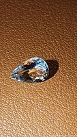 Close-up photo of a pear-shaped natural aquamarine gemstone with light blue color and faceted cut, set against a textured brown background.