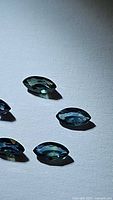 Close-up photos of five marquis cut natural sapphires with blue and green color tones, laid out on a white surface under bright light showing facets and clarity.