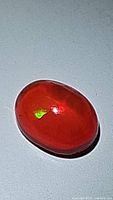 Photo showing the deep bright orange-red oval cabochon fire opal with visible green flash on its surface against a white background.