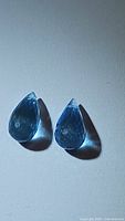 Two blue teardrop-shaped fancy briolette cut topaz gemstones shown on a light surface, showcasing the front faceted side and drilled holes for jewelry making.
