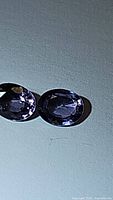 Pair of oval cut purple spinel gemstones shown on a light surface under natural and artificial light, highlighting their color and clarity.