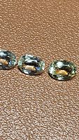 Three natural green sillimanite oval-cut gemstones arranged horizontally on a textured brown surface.