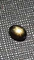 Oval cabochon cut black star sapphire gemstone placed on textured surface, showing golden star asterism under direct light.