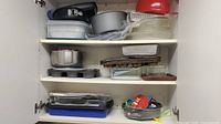 Full view of kitchen cabinet shelf filled with various bakeware including glass and metal baking sheets, springform pans, and strainers.