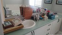 Countertop with sink showing two woven wicker baskets, assorted cleaning supplies, ceramic bathroom accessories, framed prints stacked and leaning near the wall, and a white digital bathroom scale.