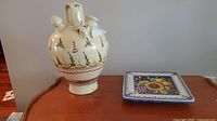Both items displayed on a wooden surface: off-white ceramic botijo jug with painted designs and a decorative wall tile with a floral pattern.