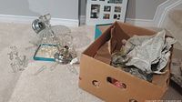 Photo showing chandelier parts including metal frame pieces and loose acrylic hanging jewels on carpet beside a cardboard box with packing paper.