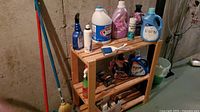 Wooden rack with multiple bottles of laundry and cleaning supplies on two shelves against a concrete wall.