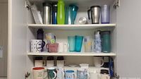 Full view of three kitchen shelves filled with assorted coffee mugs, travel mugs, plastic tumblers, and thermoses.