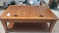 Front angled view of the wooden coffee table showing top with two removable panels and lower shelf. Surfaces show some scratches and wear.