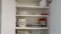Shelf with stacked white ceramic bowls and plates, multi-colored small bowls, and some clear glasses on bottom shelf.