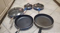 Five fry pans on tiled floor showing variety of sizes and styles, including three with lids and two without lids.
