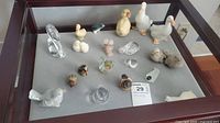 Display of multiple glass and porcelain bird figurines including ducks, geese, and an onyx burro on a grey surface inside a glass case.