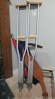 Pair of adjustable aluminum crutches leaning against wall with beige arm and hand grips. Brown leather weightlifting belt and two small hand weights visible behind crutches.