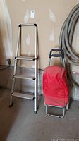 Photo of aluminum step ladder and red grocery pull cart side-by-side on concrete floor along a wall with attached large hoses.