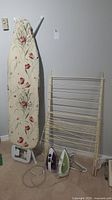 Full view of laundry care items including ironing board with floral cover, wooden drying rack, and two irons on the floor beneath.