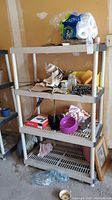 Front view of the plastic shelving unit with four shelves, showing various items placed on shelving. Shelving unit is beige/off-white and stands alone in a garage space.