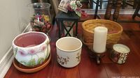 Overview of ceramic pots, black hurricane lantern, woven basket, and several candle holders with candles arranged on wooden floor.
