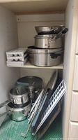 Stacked stainless steel pots and pans stored in kitchen cabinet, includes several sizes and a steamer insert, plus ice cube trays.