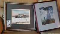 Photo showing both framed prints side by side leaning against a beige couch, one with a winter barn scene and the other with a woman and child in a field.