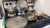 Wide angle photo of silverplate items on a kitchen stove, showing a large engraved tray, goblets, covered dishes, and other silver items