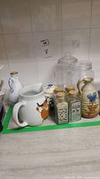 Overall view of the brown vintage Arabia milk pitcher alongside assorted ceramic and glass jars with cork lids placed on a kitchen counter.