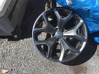 Photo showing a dark multi-spoke 20 inch Dodge rim resting on gravel near a white object and a blue tarp.