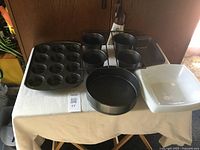 Front view of baking items including muffin pan, round springform pan, square baking dish, rectangular baking pan, and four black baking cups on stand.