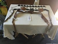 Three handmade belts laid out on a table. Two belts with metal chains, medallions, and coin charms, and one wide brown leather belt with fringed ends and stitched pattern.