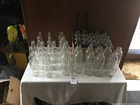 Photo showing lots of clear glass bottles arranged on a table in two sizes, one group taller and slender, the other shorter and wider, all bottles are transparent and empty.