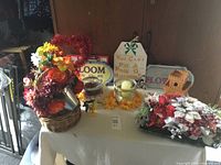 Overview of flower centerpieces and plaques arranged on a table covered with a white tablecloth, showing various floral arrangements and plaques including a tiered birdhouse plaque and inspirational quotes.