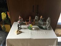 Photo showing eight collectible items on white tablecloth including Blue Mountain Pottery mermaid candle holders, floral decorated vase, glass orb paperweight, metal bird-like figurine, Inuit-style hunter figurine, metal lantern figurine, and beige carved bird figurine.