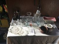 Table with various glass and ceramic collectible items displayed, including flower-shaped dish, bottles, glasses and ceramic pots.