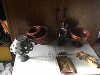 Metal tea light holder with heart-shaped cutouts, wicker basket filled partially with items, wooden pedestal bowl, female figurine with umbrella, additional figurine, framed floral artwork, and a small decorative item on a table against a wooden cabinet.