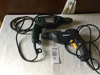 Photo showing two corded drills, one green Makita and one black and yellow Mastercraft, lying on a white cloth surface.