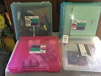 Four closed plastic storage bins in pastel colors containing assorted craft supplies visible through translucent lids.