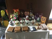 Full view of table with crafts items including glass globes containing fish figurines, vases, flower arrangements, wooden boxes, books, and small decorative items placed on a white tablecloth.
