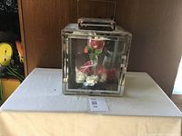 Front view of a square glass and metal curio box on a table, showing artificial red and white flowers inside.