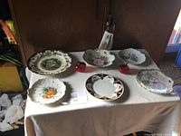 Set of six decorative plates and two red glass apple figurines arranged on a table with a white cloth against a wood background