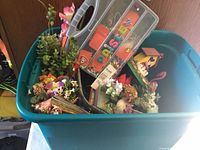 View of plastic bin filled with artificial flowers, small floral arrangements, birdhouse and a decorative prayer box.