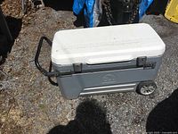 Large gray and white rolling cooler by Igloo with wheels, handle, and metal lid latches. Cooler lid has a ruler on top. Cooler is outdoors on gravel ground showing used condition.