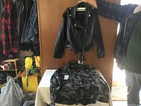 Photo showing the black leather jacket hanging with gold-tone zipper and belted waist, and the grey/black camouflage hunting coat laid flat on table.