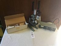 Photo showing the microscope, open box with microscope accessories, and SWR meter on a white surface with wooden background.